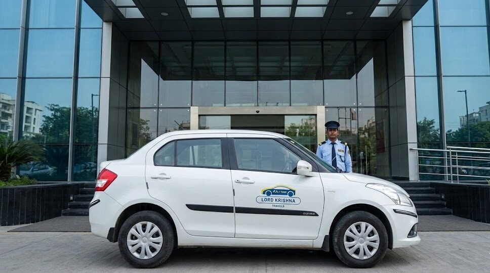 a white lord krishna travels sedan parked at a modern corporate office building in noida, with a uniformed driver waiting.